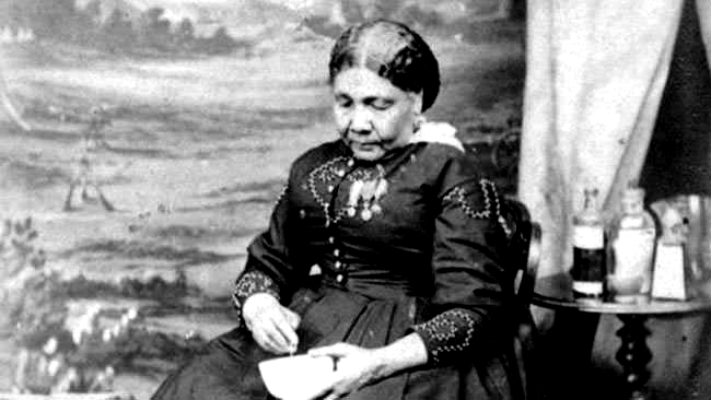 Photographie en noir et blanc de Mary Seacole, infirmière et humanitaire jamaïcaine-britannique du XIXe siècle, vêtue d'une robe victorienne sombre ornée de broderies décoratives. Elle est assise, tenant un bol, des flacons de médicaments posés sur une table voisine, symbolisant sa contribution à la médecine sur le champ de bataille pendant la guerre de Crimée.